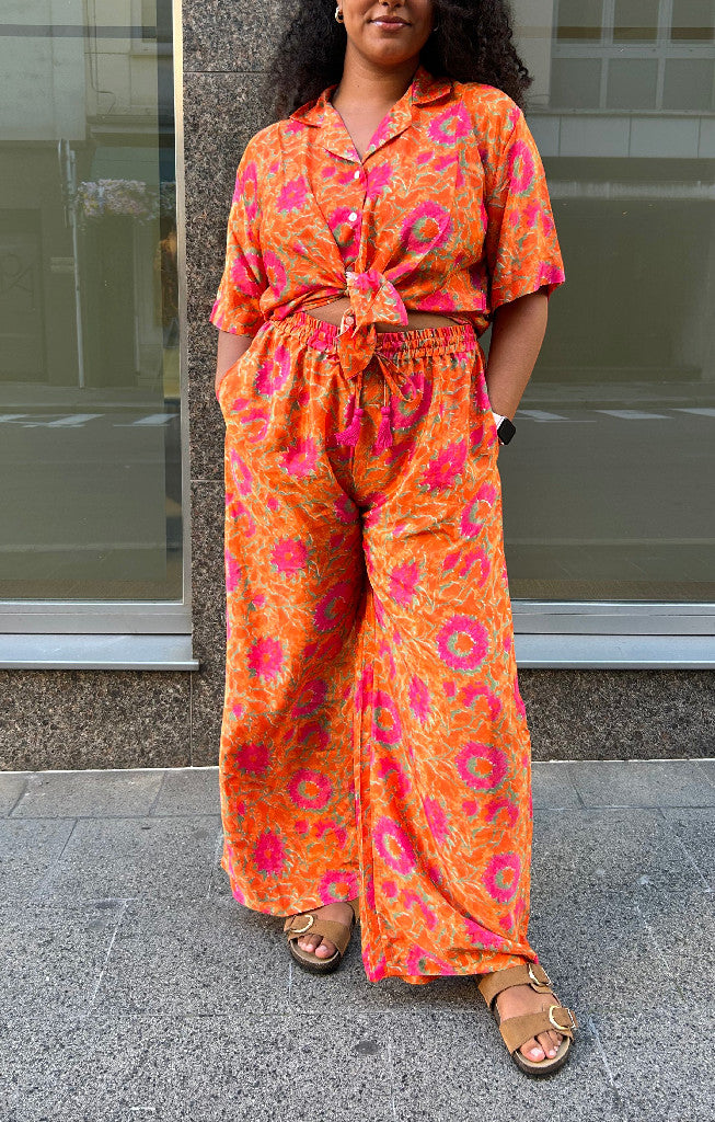 Ensemble Devï Studios SARL deux pièces en soie et viscose orange à imprimé floral rose et vert, chemise courte nouée et pantalon large.